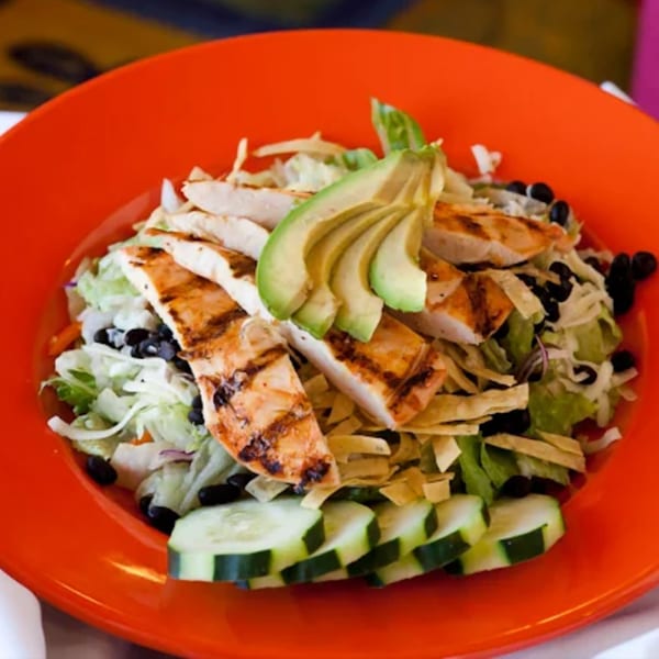 Grilled chicken salad with avocado, black beans, tortilla strips, and cucumbers in an orange bowl.
